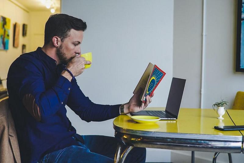 Man sitting at a desk reading a book and drinking a cup of tea